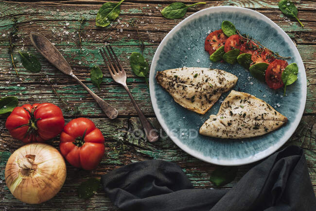 Von oben gebratener Fisch mit Basilikum auf schäbigem Holztisch neben frischen Tomaten und Zwiebeln — Stockfoto