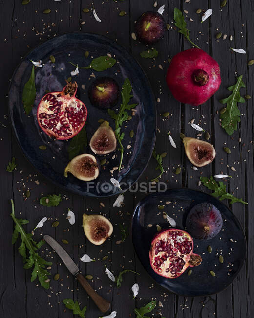 Vista superior de figos cortados frescos e romãs colocados em placas perto de arugula e faca na mesa de madeira — Fotografia de Stock