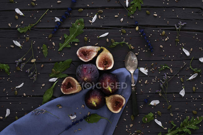 From above fresh halved figs and silver spoon placed on wooden table near blue napkin and arugula in kitchen — Stock Photo