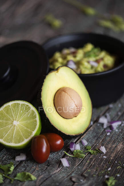 Fresh lime and tomatoes placed near halved avocado and pot with guacamole on lumber table — Stock Photo