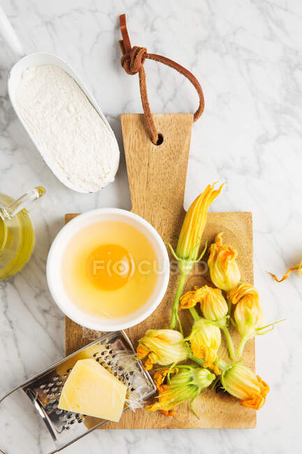 From above grater with cheese and bowl with raw egg placed on cutting board near fresh zucchini flowers and flour on marble table in kitchen — Stock Photo