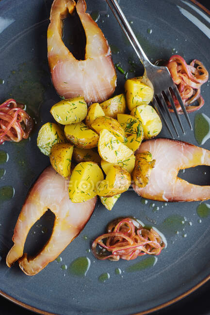 De arriba trozos de pescado marinado y cebollas con patatas asadas dispuestas en plato cerca de tenedor - foto de stock