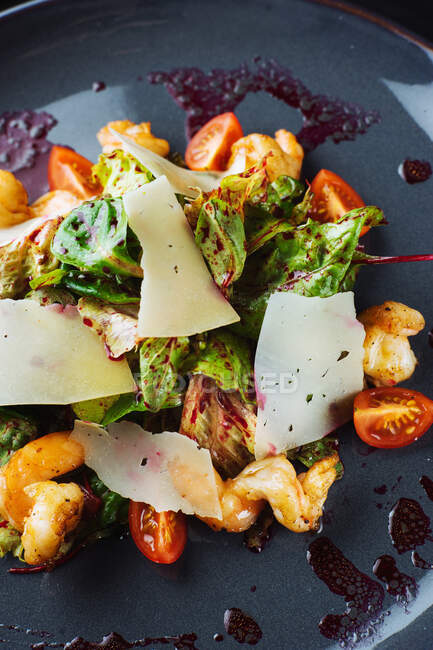 Зверху крупним планом смачний салат зі свіжими овочами та листям з креветками та сиром, поданий на чорній тарілці з соусом — стокове фото