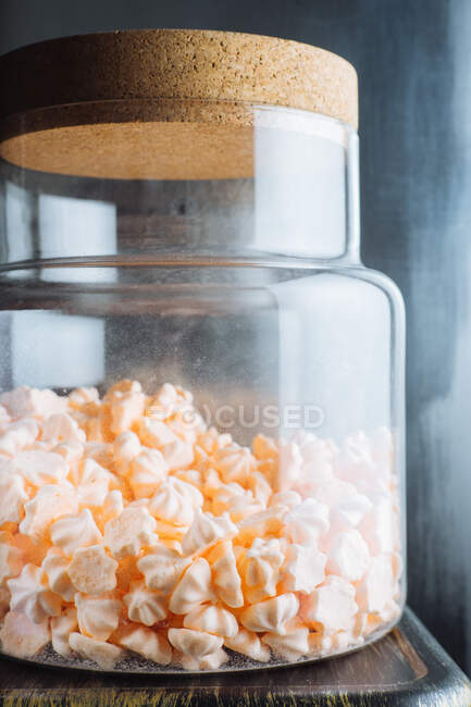 Sweet peach colored mini meringues in big glass jar placed on table next to window — Stock Photo