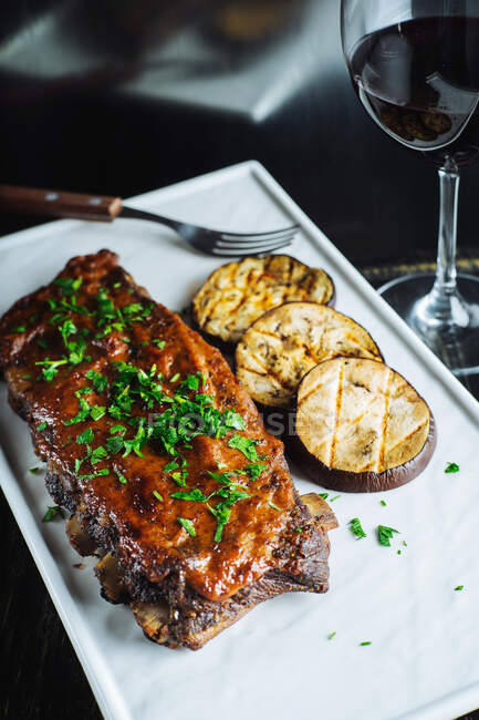 Top view of delicious grilled pork ribs garnished with fresh parsley served with sliced grilled eggplant on white tray — Stock Photo