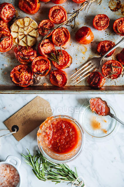 Top view of glass pot with homemade tomato sauce placed on table next to metal tray with grilled tomatoes with garlic and rosemary — Stock Photo