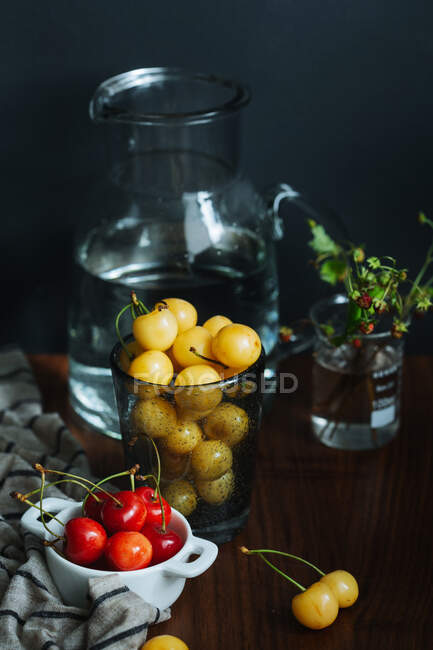 Свежие красные и желтые плоды вишни в стеклянных кастрюлях на деревянном столе возле стеклянной вазы с водой на черном фоне — стоковое фото