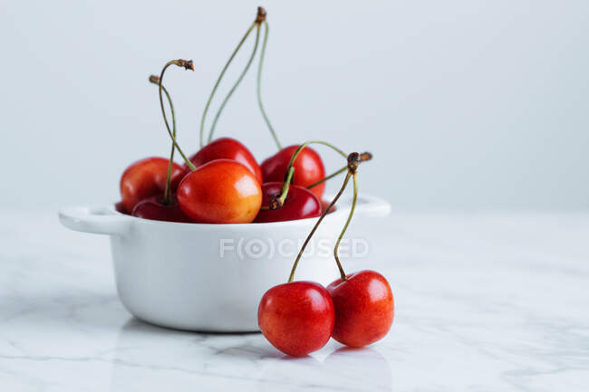 Cerise rouge mûre fraîche avec des tiges dans un pot en céramique blanche placé sur une table en marbre contre un mur blanc — Photo de stock