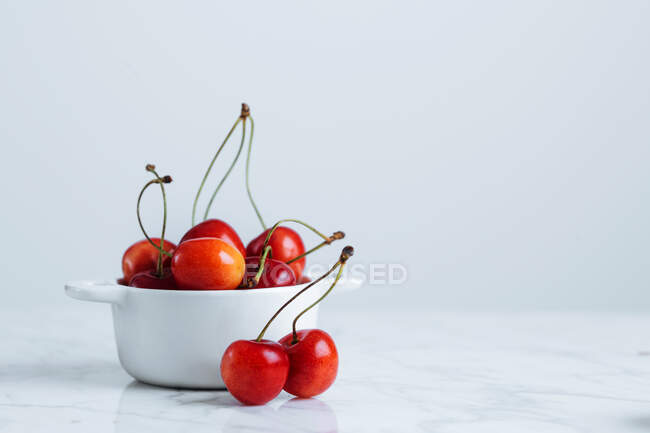 Frische reife rote Kirsche mit Stielen in weißem Keramiktopf auf Marmortisch vor weißer Wand — Stockfoto