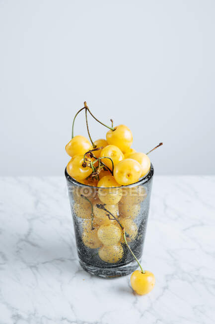 D'en haut de verre plein de cerises jaunes fraîches avec des tiges sur la table de marbre sur fond blanc — Photo de stock