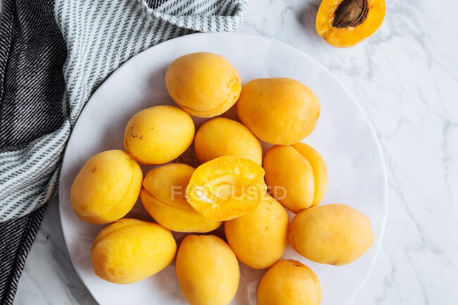 Вид зверху на білу тарілку зі свіжими жовтими стиглими абрикосами, розміщеними на тарілці біля столової тканини на білому мармуровому столі з нарізаним навпіл абрикосом — стокове фото