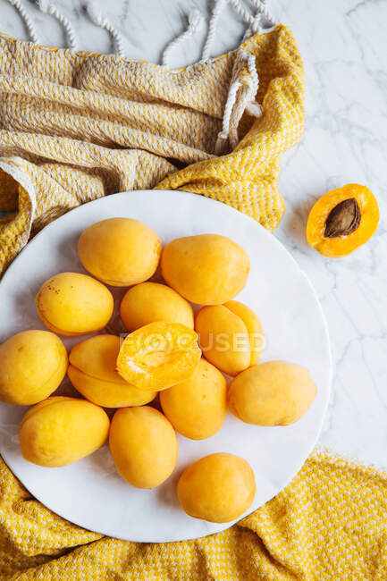 Draufsicht auf weißen Teller mit frischen gelben reifen Aprikosen auf gelbem Tuch auf weißem Marmortisch mit halbierter Aprikose — Stockfoto