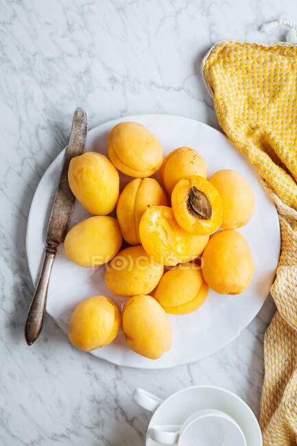 Вид зверху на білу тарілку зі свіжими жовтими стиглими абрикосами, розміщеними на жовтій тканині на білому мармуровому столі з нарізаним навпіл абрикосом — стокове фото