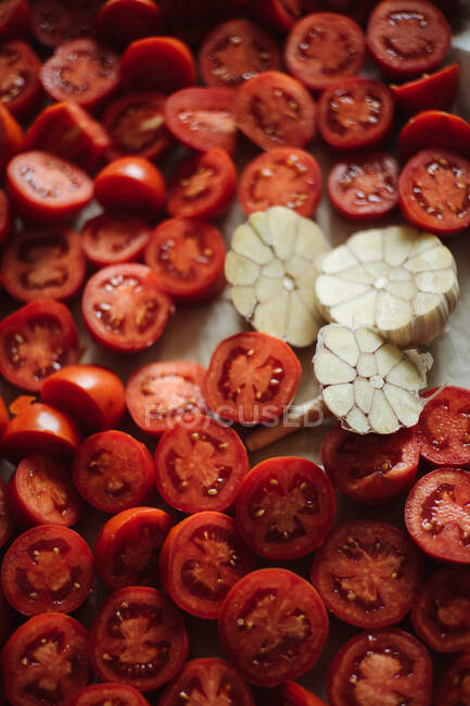 Вид сверху свежих спелых красных помидоров вишни, разрезанных пополам, и чеснока, приготовленного для варки. — стоковое фото