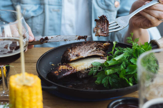 Cultivez une personne méconnaissable avec un couteau et une fourchette en mangeant un délicieux steak de bœuf grillé avec des os servi avec de la verdure fraîche et du maïs grillé au restaurant — Photo de stock