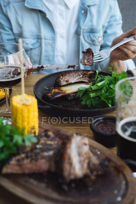 Crop unrecognizable person with knife and fork eating delicious grilled beef steak with bone served with fresh greenery and grilled corn in restaurant — Stock Photo