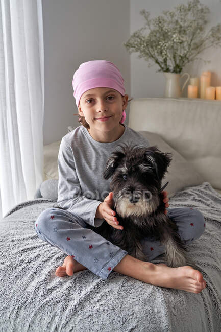 D'en haut adorable petit garçon malade en bandana rose et pyjama caressant animal de compagnie assis sur le lit à la maison en regardant la caméra — Photo de stock
