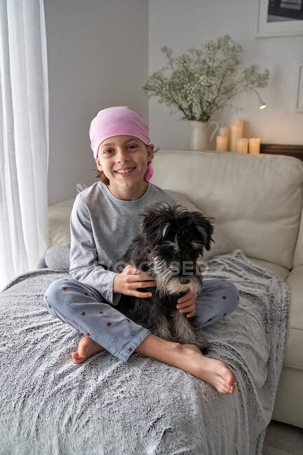 From above adorable sick little boy in pink bandana and pajama caressing pet while sitting on bed at home looking at camera — Stock Photo