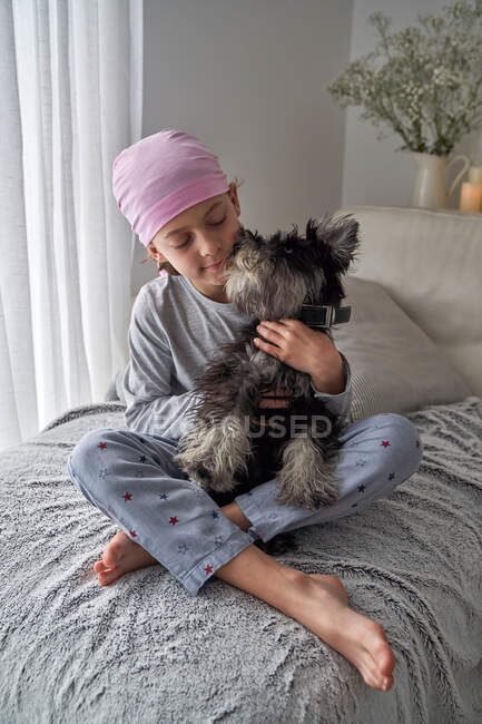 De cima adorável menino doente em bandana rosa e pijama acariciando animal de estimação enquanto sentado na cama em casa — Fotografia de Stock