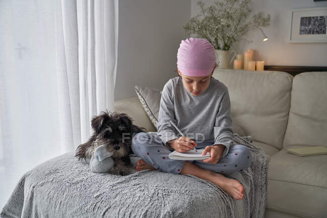Dal basso bambino felice con malattia del cancro annotazioni di scrittura mentre seduto con cane sul letto in camera — Foto stock