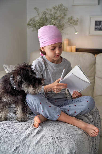 Знизу щаслива дитина з раковими захворюваннями пише нотатки, сидячи з собакою на ліжку в кімнаті. — стокове фото
