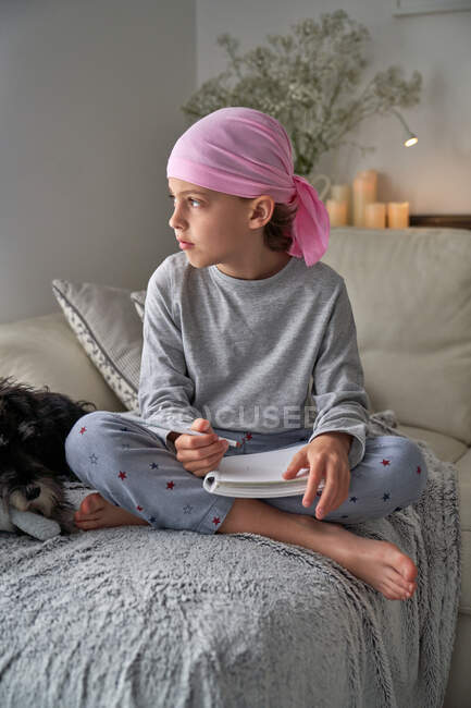 Знизу серйозна дитина з діагнозом раку робить нотатки, сидячи на ліжку в кімнаті — стокове фото