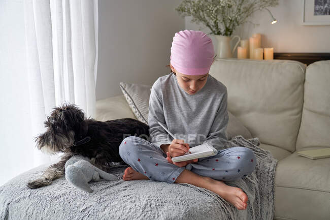 Знизу щаслива дитина з раковими захворюваннями пише нотатки, сидячи з собакою на ліжку в кімнаті. — стокове фото