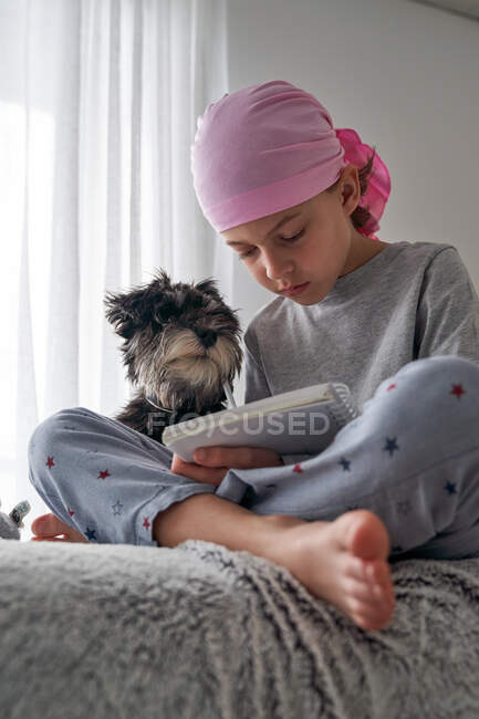 Desde abajo niño feliz con cáncer escribir notas mientras está sentado con el perro en la cama en la habitación - foto de stock