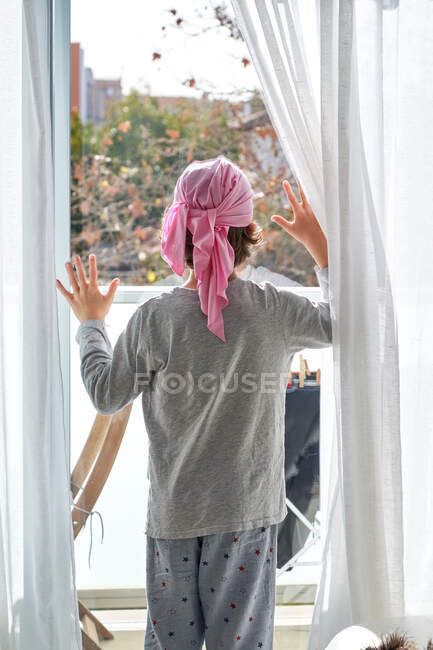 Visão traseira de criança anônima com câncer vestindo bandana rosa e colocando as mãos na janela no quarto — Fotografia de Stock