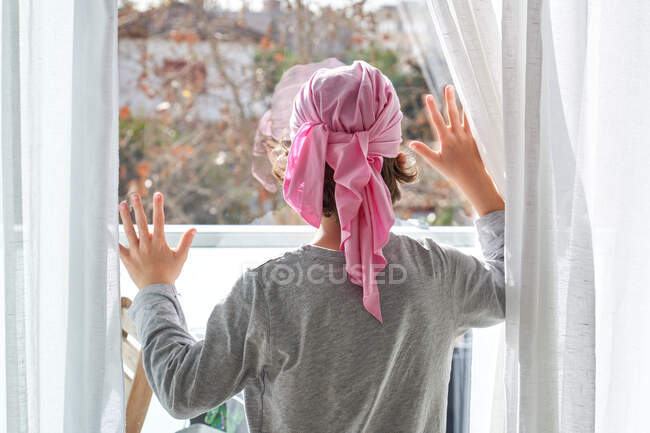 Visão traseira de criança anônima com câncer vestindo bandana rosa e colocando as mãos na janela no quarto — Fotografia de Stock