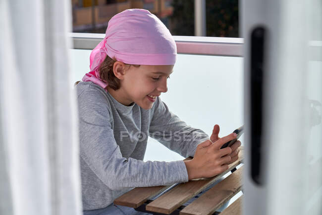Niño alegre con enfermedad del cáncer disfrutando de pasatiempo con teléfono celular en la terraza - foto de stock
