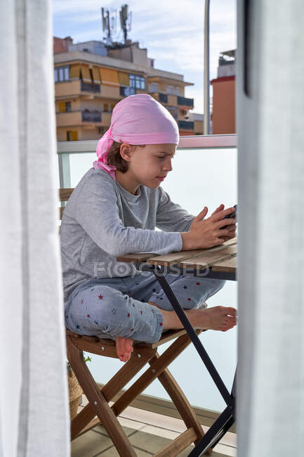 Niño alegre con enfermedad del cáncer disfrutando de pasatiempo con teléfono celular en la terraza - foto de stock