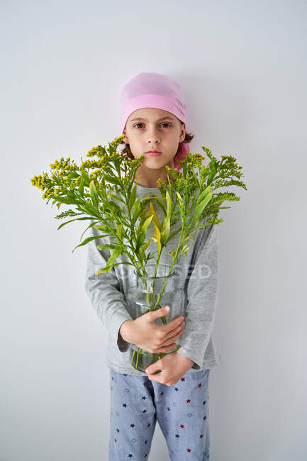 Сосредоточен маленький мальчик с диагнозом рак носить розовую бандану и смотреть в камеру, держа вазу с цветами и стоя у стены — стоковое фото