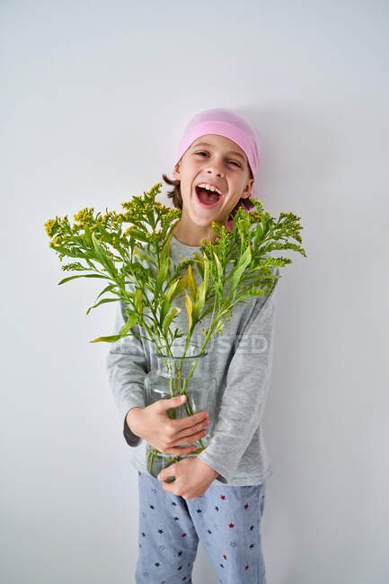 Веселий хлопчик з діагнозом раку в рожевій бандані і дивиться на камеру, тримаючи вазу з квітами і стоячи біля стіни. — стокове фото
