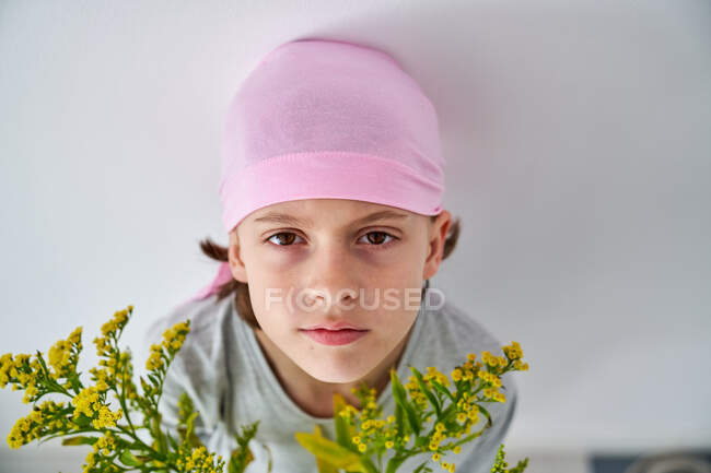 Niño enfocado con diagnóstico de cáncer usando bandana rosa y mirando a la cámara mientras sostiene el jarrón con flores y de pie en la pared - foto de stock