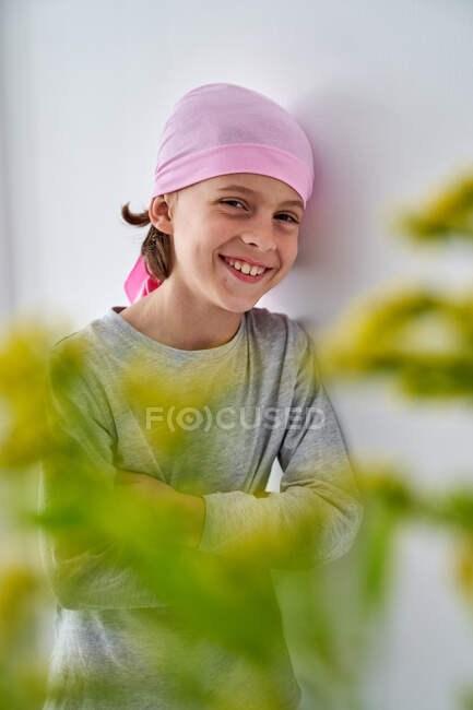 Веселий хлопчик з діагнозом раку в рожевій бандані і дивиться на камеру, тримаючи вазу з квітами і стоячи біля стіни. — стокове фото