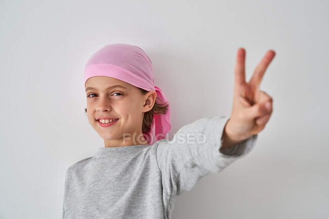 Brave small child with cancer diagnosis looking at camera making victory gesture with fingers on gray background — Stock Photo