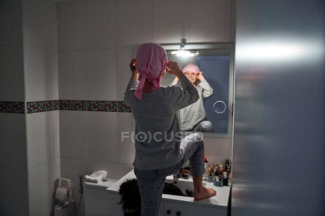 Visão traseira da criança doente colocando bandana rosa na frente do espelho no banheiro — Fotografia de Stock