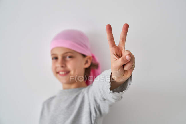 Petit enfant courageux avec diagnostic de cancer en regardant la caméra faire un geste de victoire avec les doigts sur fond gris — Photo de stock