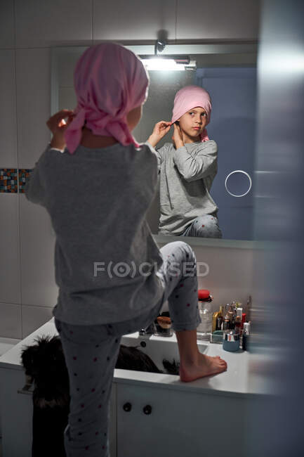 Вид сзади больного ребенка, надевающего розовую бандану перед зеркалом в ванной комнате — стоковое фото