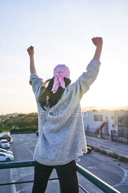 Vue arrière de la femme en bandana rose levant les mains et contemplant le coucher du soleil tout en se tenant sur le toit de l'immeuble en ville — Photo de stock