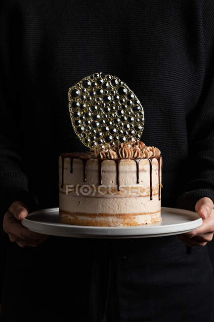 Unrecognizable person holding a festive cake with chocolate filling and frosting and silver isomalt decoration on table with black background — Stock Photo