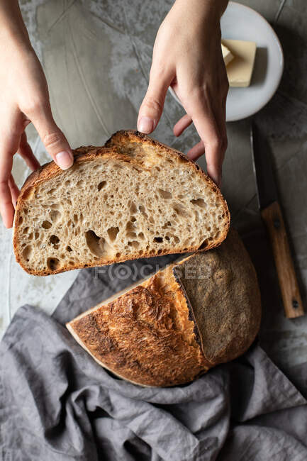 De arriba de la cosecha hembra sosteniendo cortar la mitad de deliciosa masa fermentada crujiente pan pan sobre mesa de madera con cuchillo y mantequilla - foto de stock