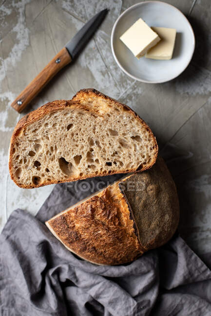 Corte la mitad del delicioso pan crujiente de masa madre sobre una mesa de madera con cuchillo y mantequilla - foto de stock