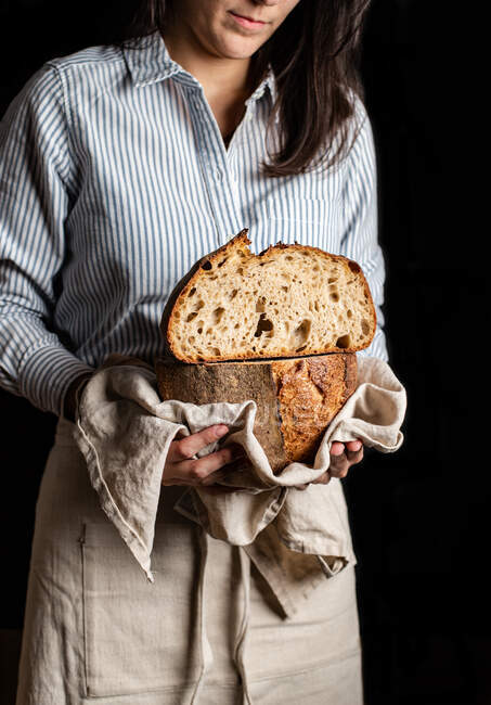 Mujer de la cosecha panadero en delantal celebración de corte en media hogaza de pan fresco y saludable artesanal - foto de stock