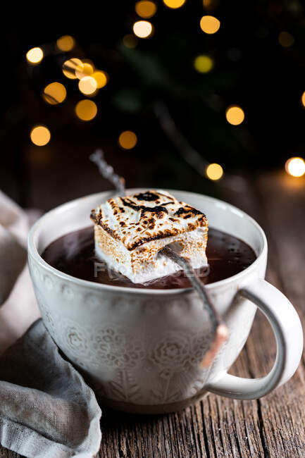 Tazza bianca con gustosa bevanda di cioccolata calda guarnita con marshmallow arrosto posto su tavolo di legno su sfondo scuro con scintille — Foto stock