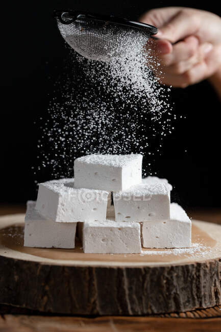 Crop personne avec tamis saupoudrer de sucre en poudre sur des morceaux de guimauve placés sur une planche de bois sur fond noir — Photo de stock