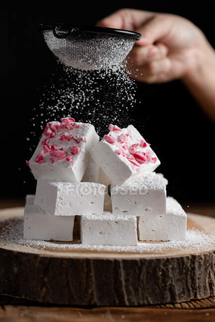 Crop person with sieve sprinkling sugar powder over pieces of marshmallow placed on wooden board against black background — Stock Photo