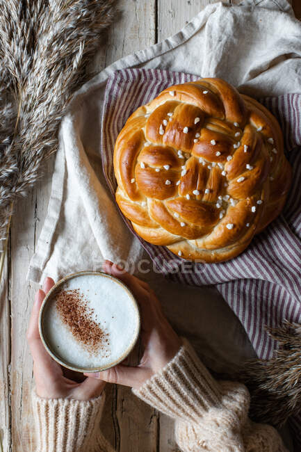 From above of crop female in beige sweater holding cup of hot cappuccino while sitting at wooden table with fresh homemade braided bread placed on fabric next to animal fur — Stock Photo