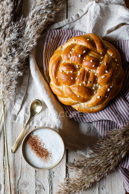 Tazza di cappuccino caldo seduto al tavolo di legno con pane fresco fatto in casa intrecciato posto su tessuto accanto alla pelliccia animale — Foto stock